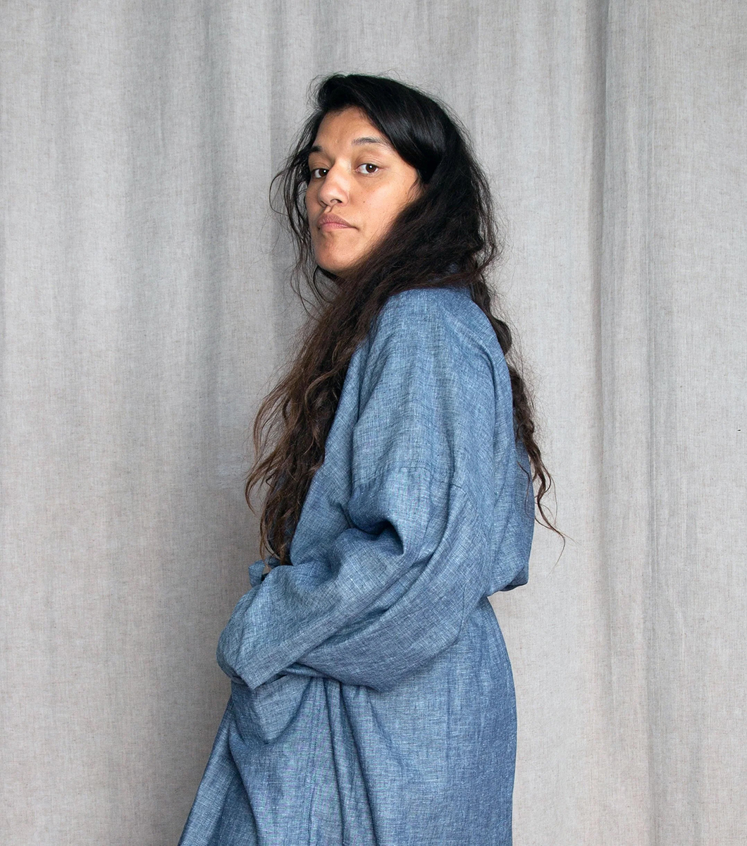 Pelenakeke, a brown-skinned woman with long wavy hair, stands slightly twisted towards the camera, looking at us. She wears a long denim coat.
