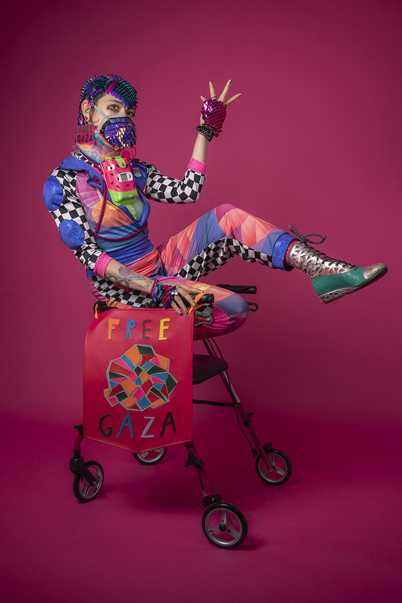 Sky, a nonbinary person with tattoos and bold makeup, poses on a hot pink rollator wearing a colorful geometric jumpsuit, sculptural accessories, and a respirator mask, with a “Free Gaza” banner attached to the rollator.