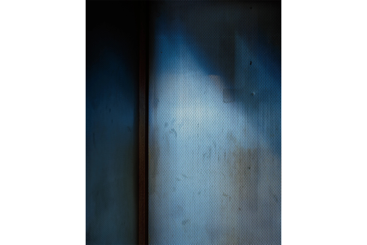 Photo of an interior room corner with the wall to the right perforated with small holes and covered in dark blue and brown shadows.
