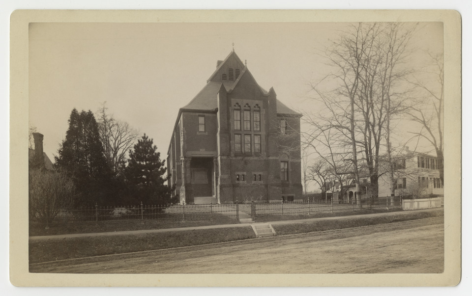 Sepia-toned photograph of a collegiate, stone building with a pointed roof. It stands behind an empty street and sidewalk, with trees and other, partially visible buildings on either side.