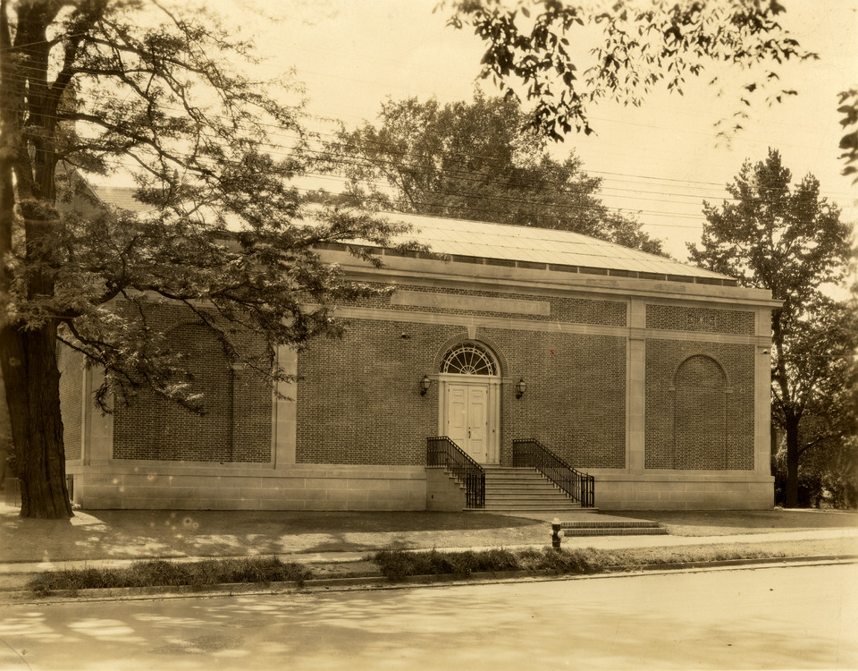 Sepia-toned photograph of a long and low brick building. It stands behind an empty street and sidewalk. Several steps lead up from the sidewalk to a pair of white doors on the front facade of the building.