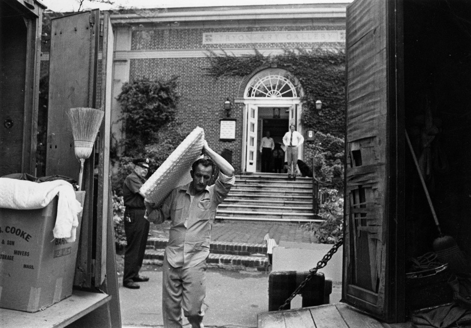 Black-and-white photograph focused on a worker carrying a large, flat rectangular package above his head. A brick building with open doors stands in the background. The open backs of two moving trucks are visible in the foreground.
