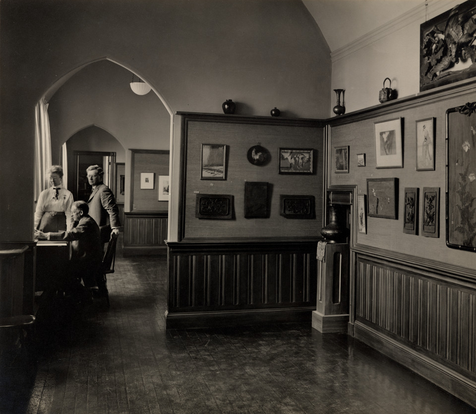 Black-and-white photograph of an art gallery. Framed artwork and small ceramic vessels are on display. Three people are grouped on the left side of the image through an arched doorway.