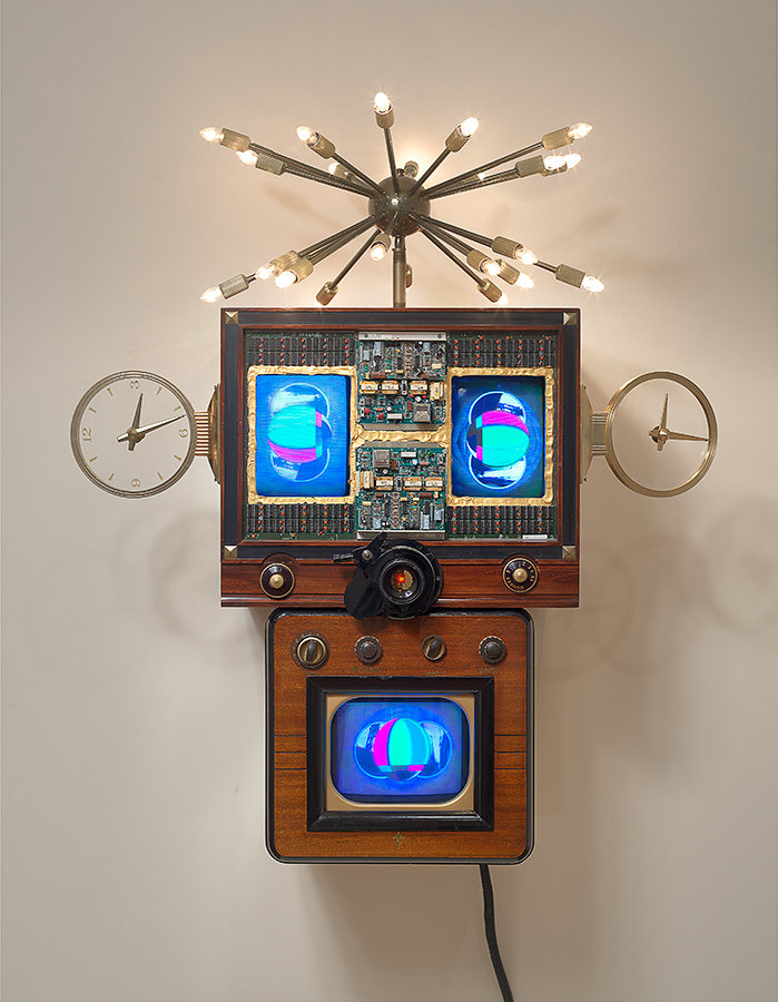 A sculpture made of various electronic materials. A television and two screens are positioned as if to form a face, with two eyes and a mouth. Clocks on either side imply ears, and a spiky light fixture on top seems like hair.