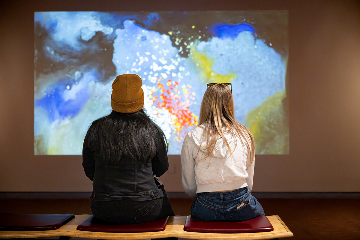 two people seated, looking at an artwork together. 