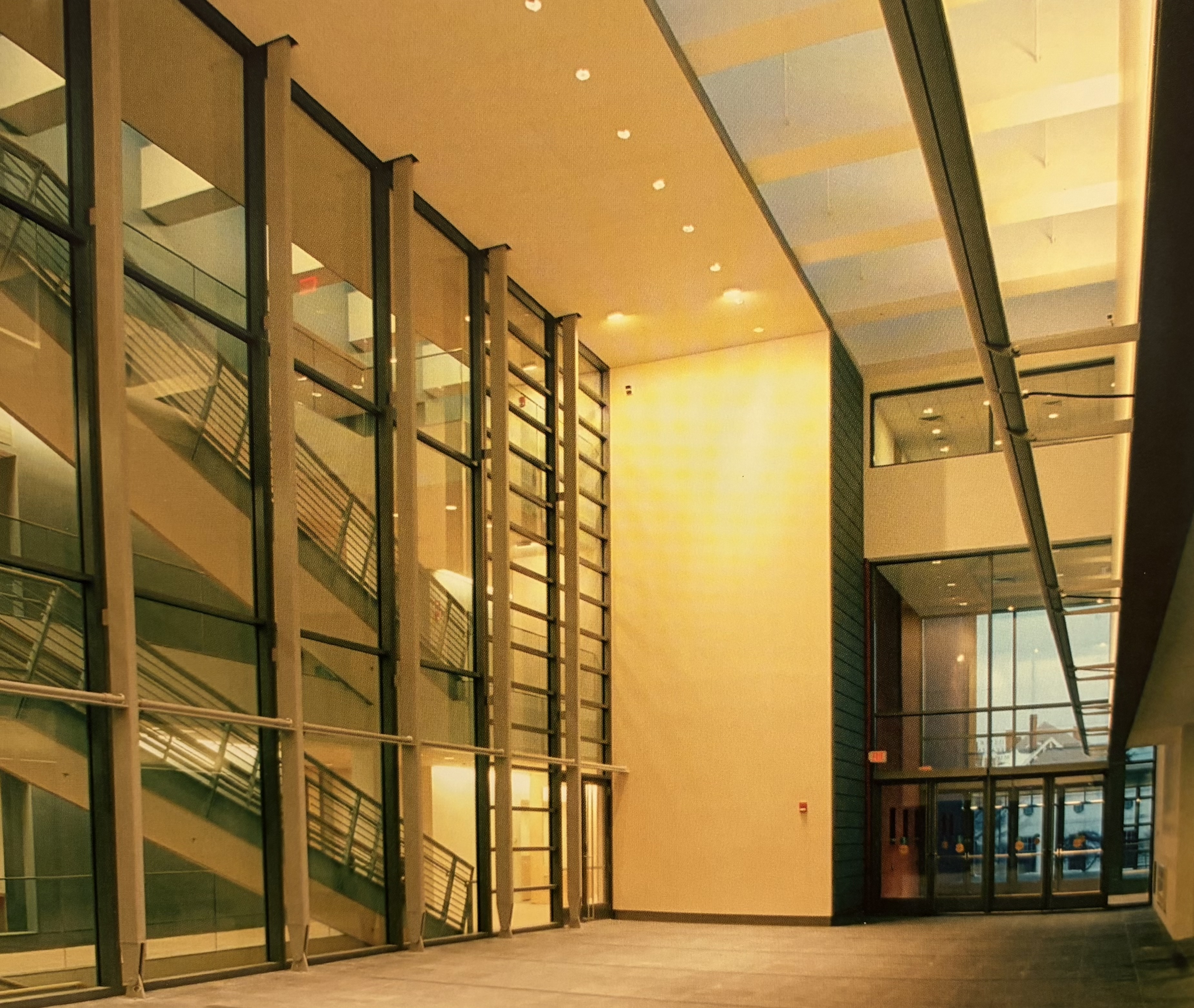Interior view of an open atrium. Large windows on the left edge of the image reveal two flights of interior staircases. Glass doors at the end of the atrium lead to the outdoors.