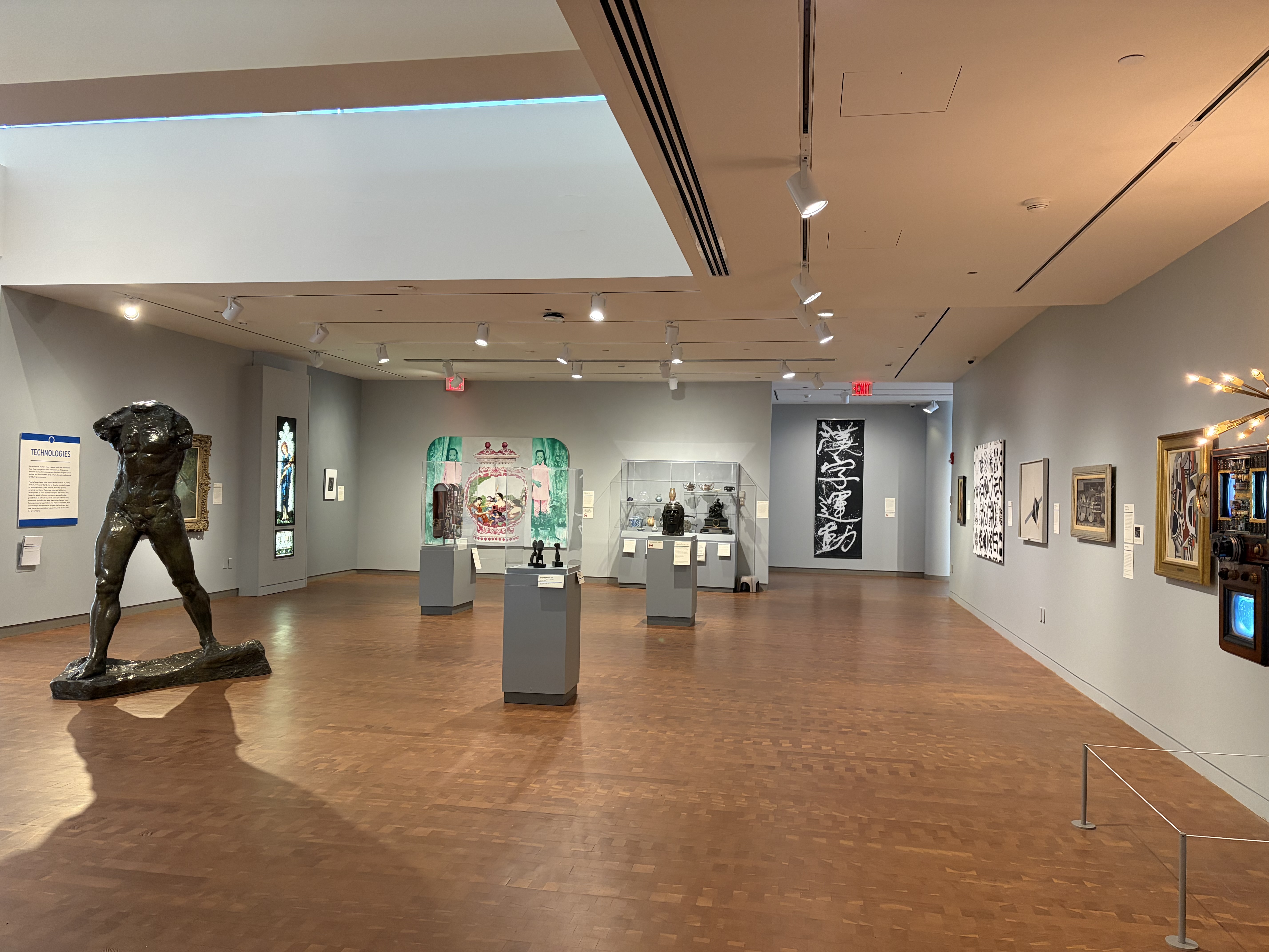 Interior view of an art gallery with light grey walls and brown, wooden floor. Various large paintings hang on the walls. Several pedestals and cases are scattered around the space.