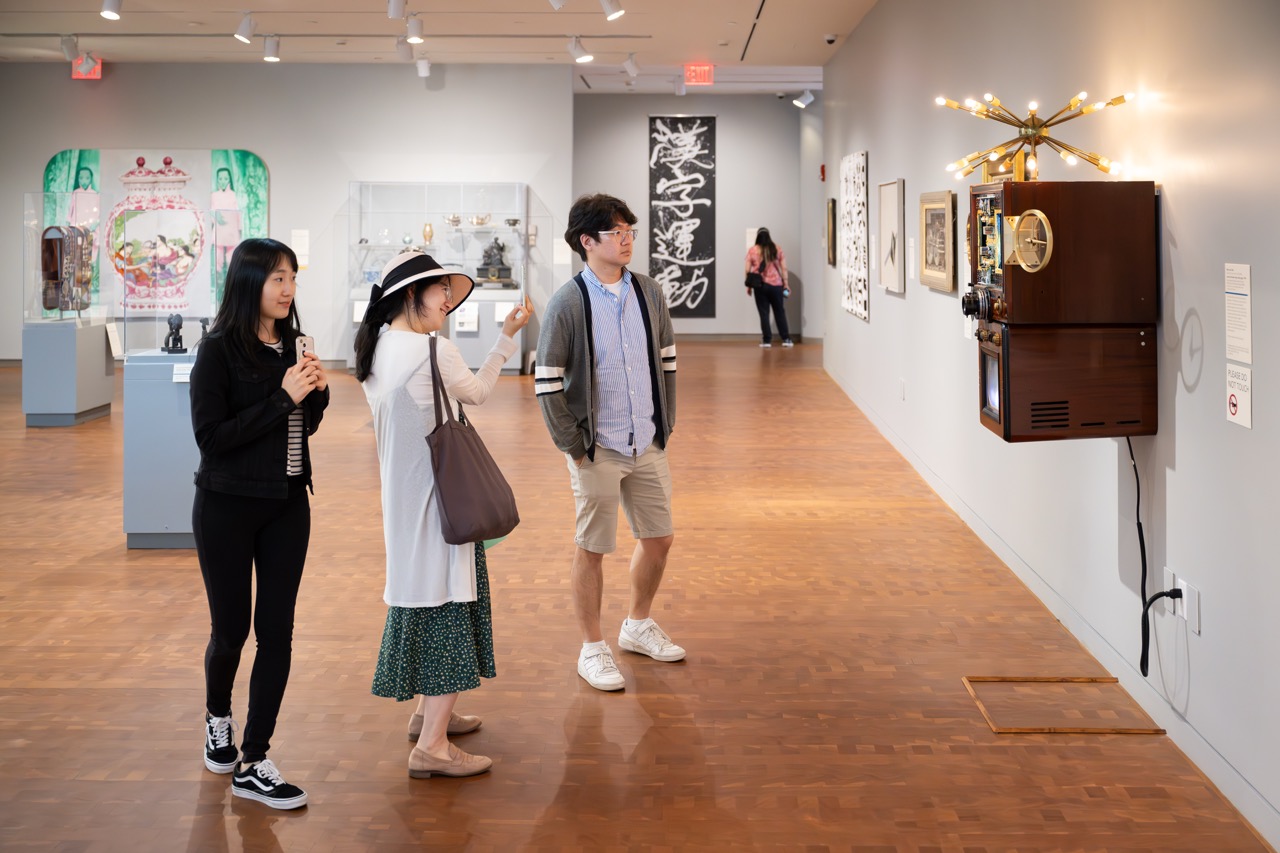 Three people engaging with a work on art on the wall by taking a photo and looking at the artwork