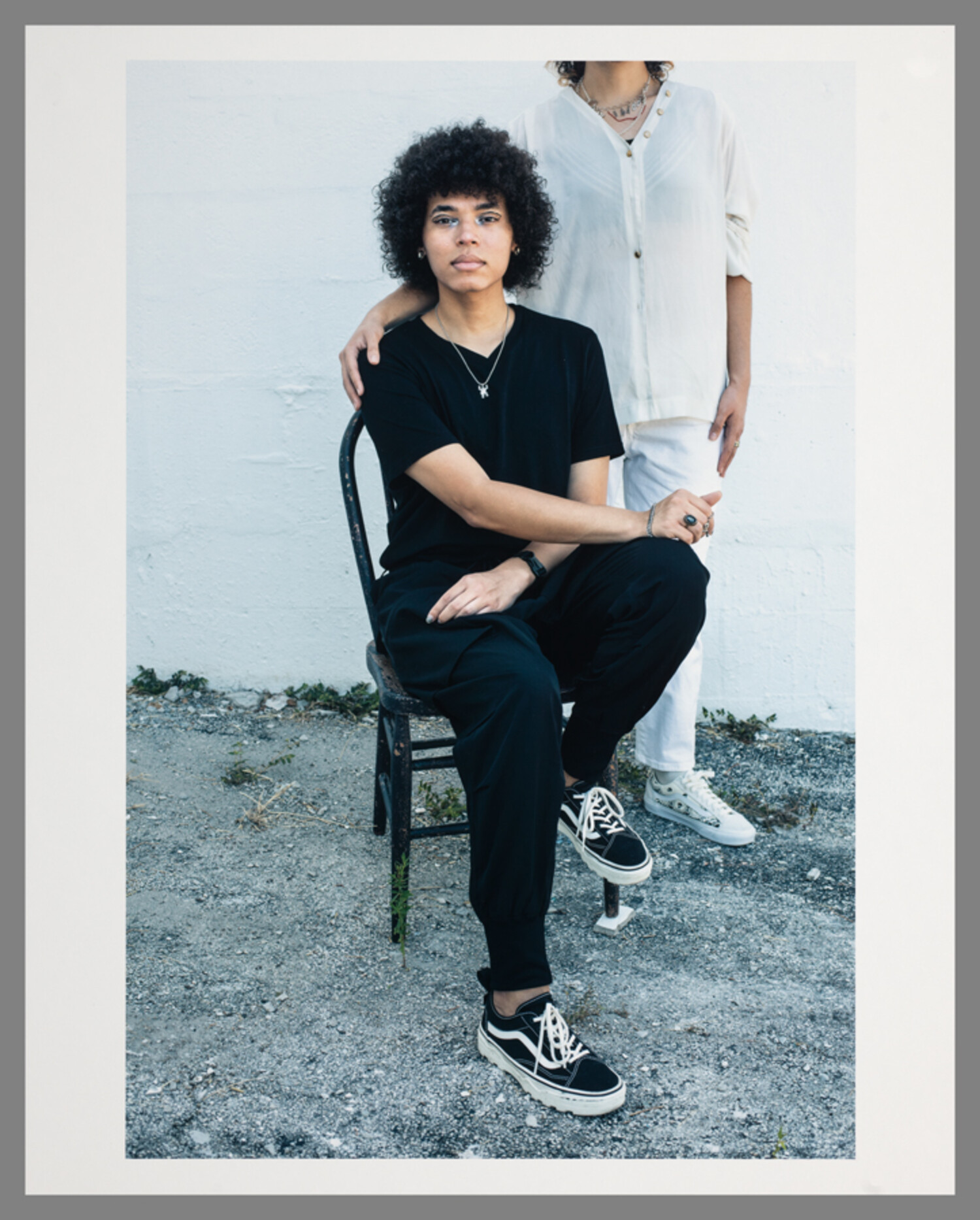 Portrait photograph centered on a person sitting in a chair. Another person stands behind them with an arm around the sitting person’ shoulders, and their head out of frame.