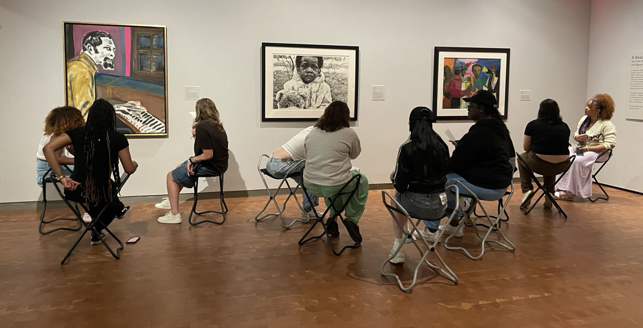 Nine people sitting on stools studying 3 paintings on the wall