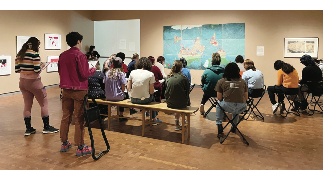 Gallery of people sitting on stools and standing looking at a map-like artwork on the wall
