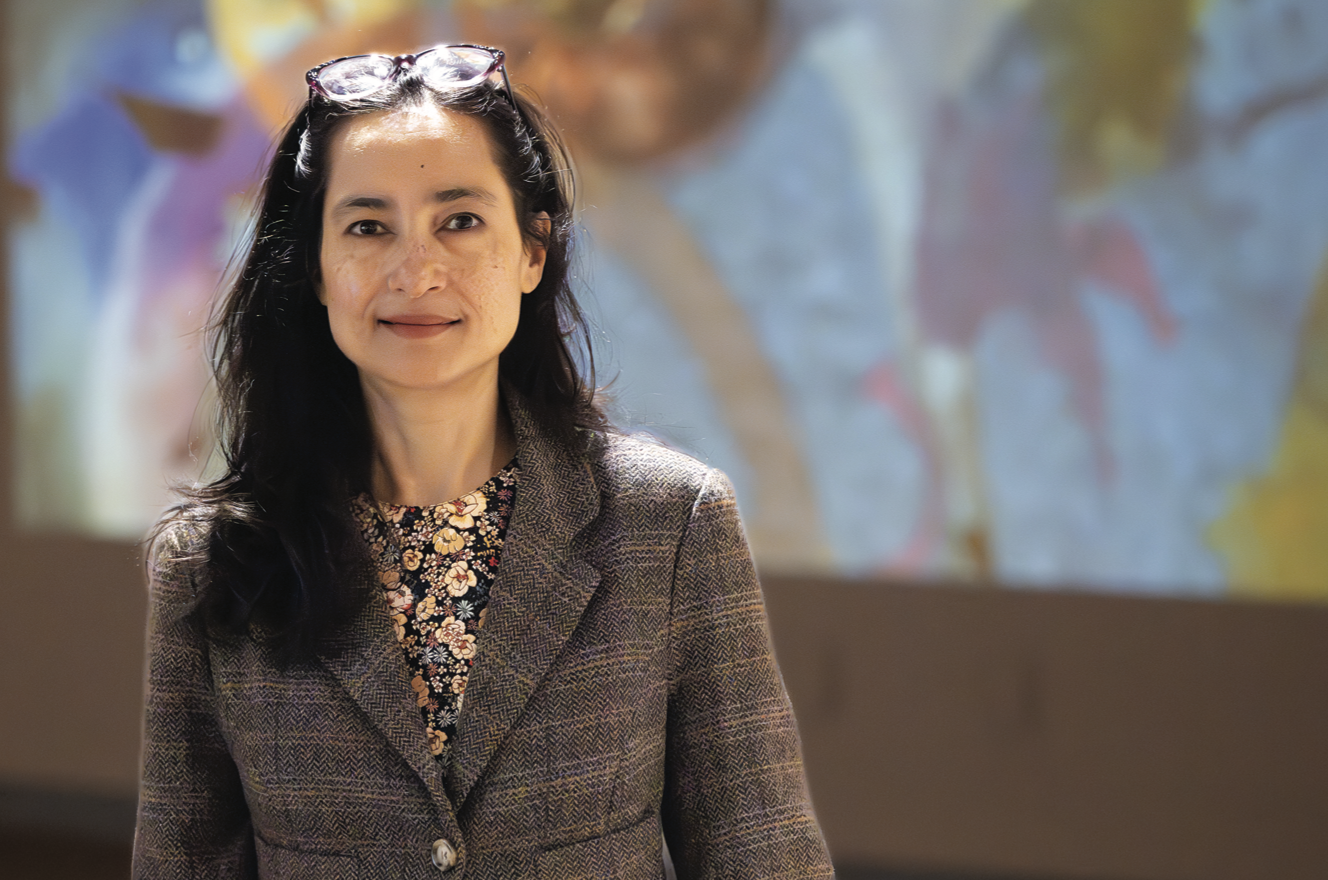 Woman with long black hair wearing a tweed jacket, stands in front of a projected image on the wall behind