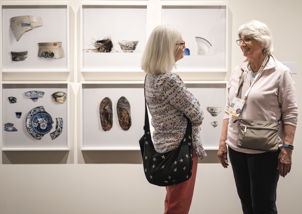 Two people with gray hair standing and talking in front of a series of photographs on the wall
