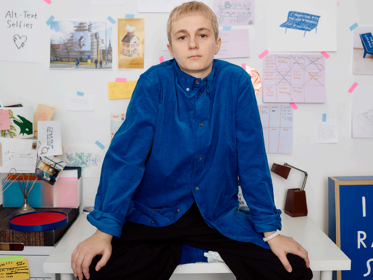 Me, a white person in a bright blue corduroy shirt and bleached blonde hair. Looking directly at the camera, relaxed but not smiling. Taped to the wall behind me are various notes about projects, sketches, and things I like. 