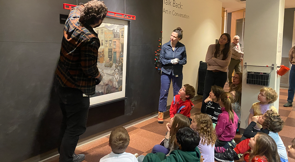 Two people demonstrating hanging an artwork to a class of young elementary students