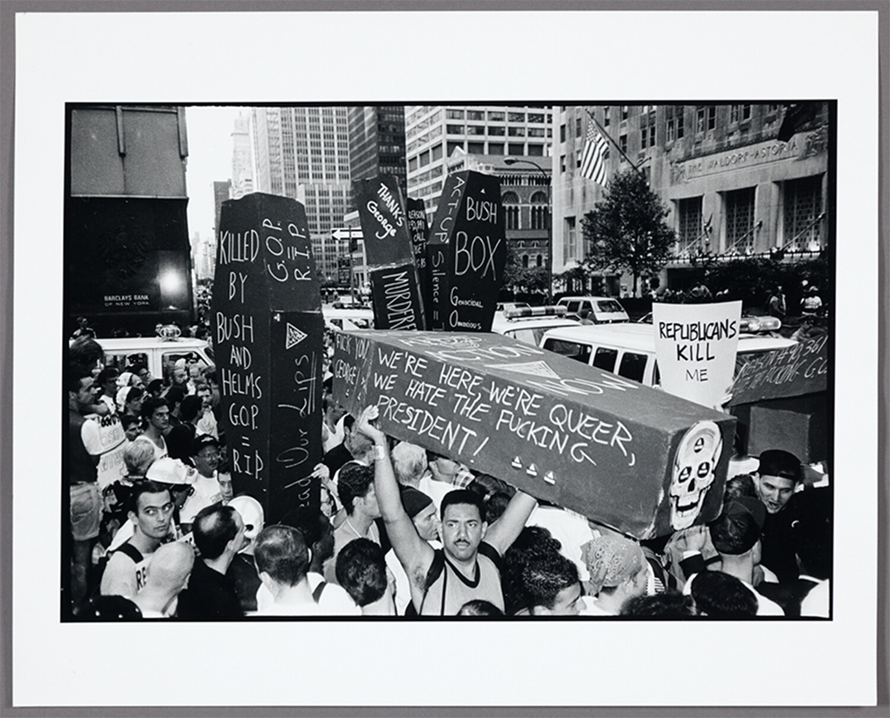 Photograph of protesters gathered on a crowded city street with cars in the background. Protest signs read, “Killed by Bush and Helms” and, “We’re here, we’re queer, we hate the fucking President!”