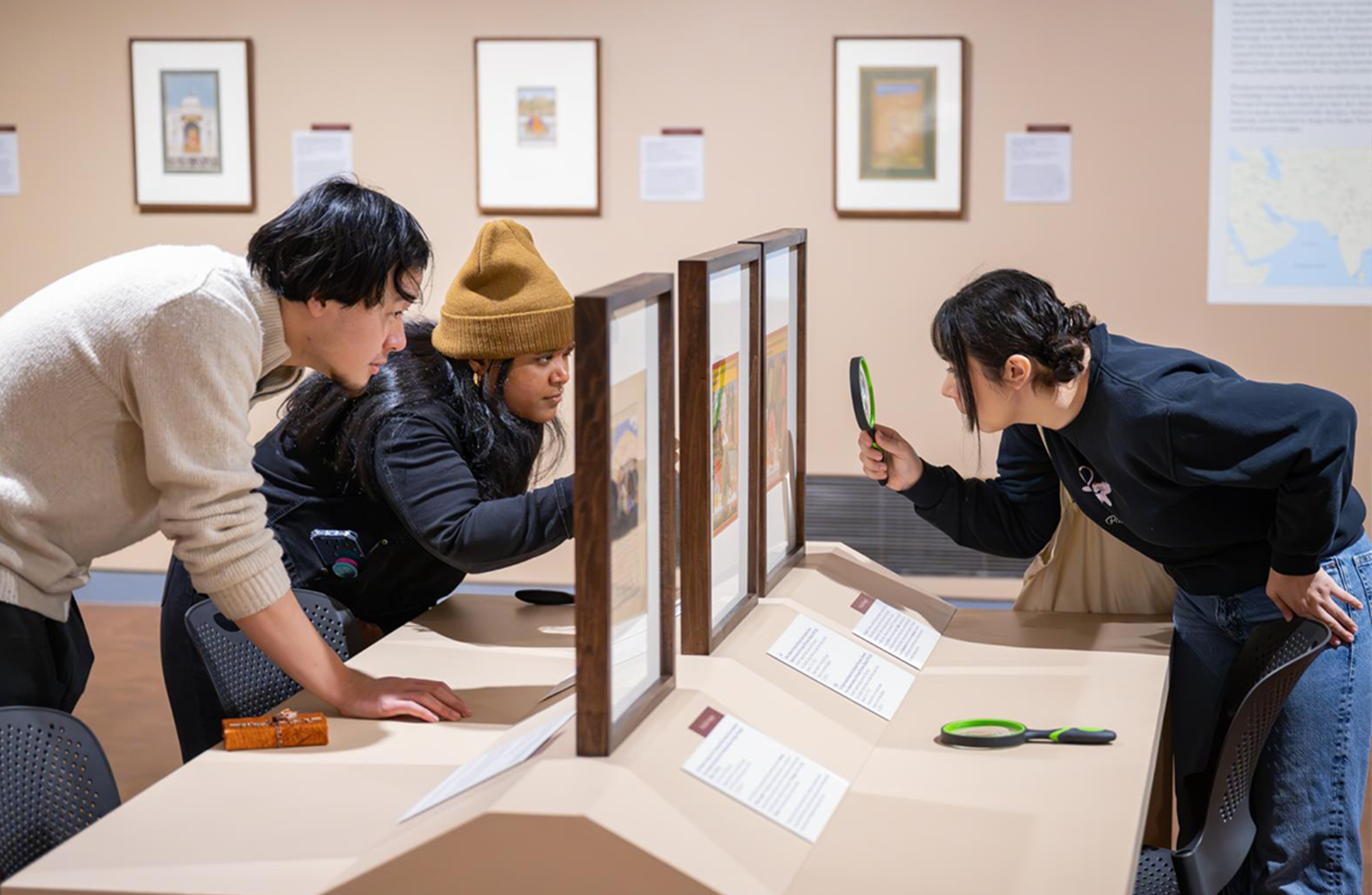 Three people in a gallery looking closely at artwork, two with magnifying glasses