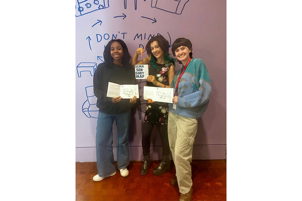 Three young people standing in front of the exhibition title wall for Don’t mind if I do, and holding final versions of the SCMA sensory map. All three of them are smiling widely.