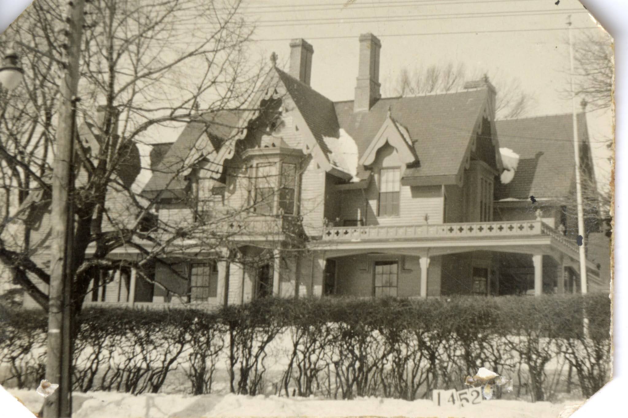 A sepia photograph of a large wood house with a wrap-around porch and two chimneys, surrounded by snow and trees.