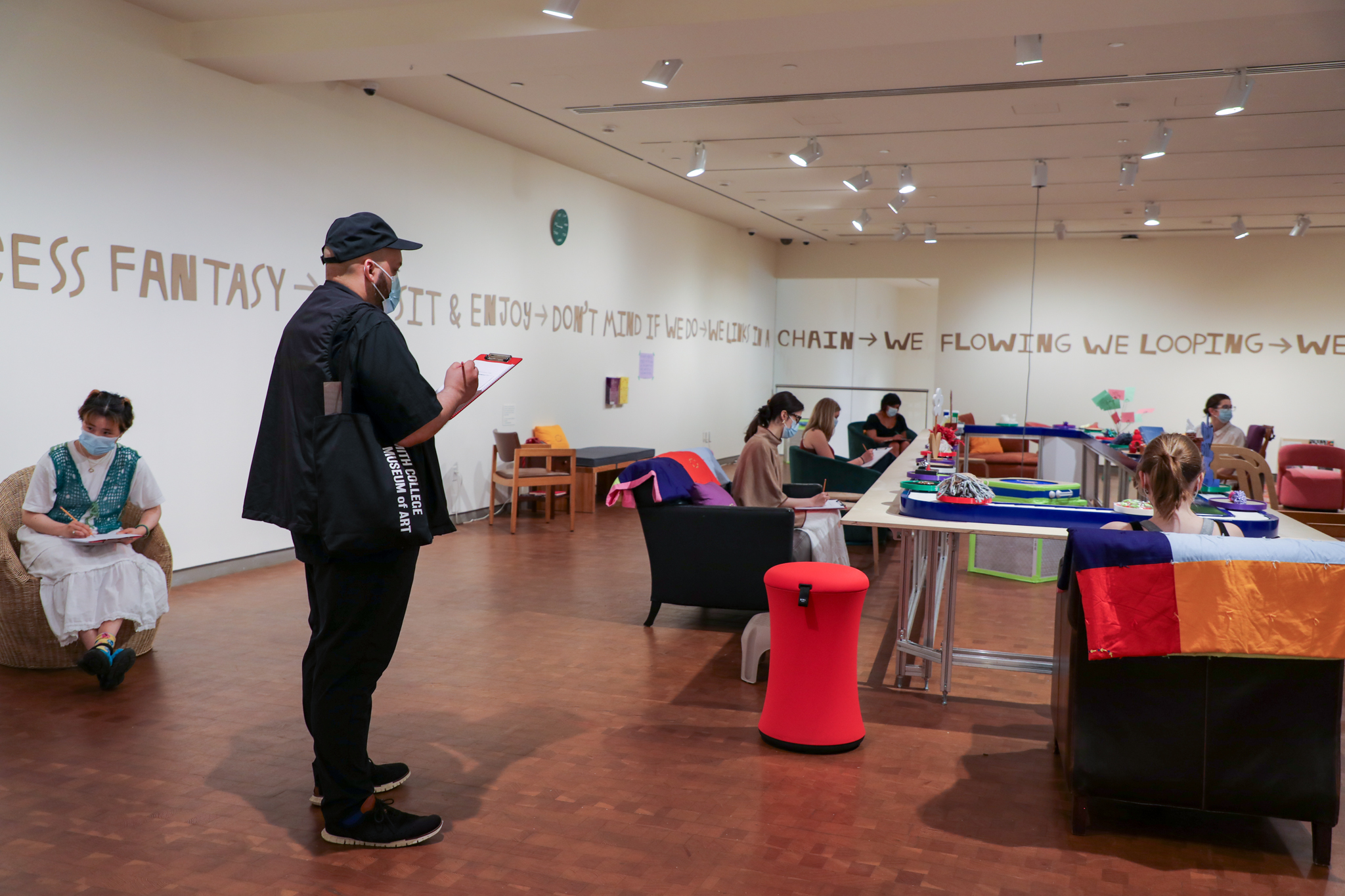Man on left with a clipboard in a gallery with conveyor belt with art on it surrounded by different forms of seating, with several people seated and wearing masks
