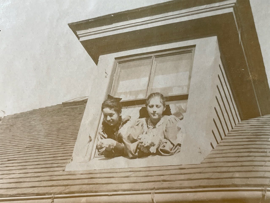 Sepia photograph of two girls with their hair pulled back leaning out of a dormer window.