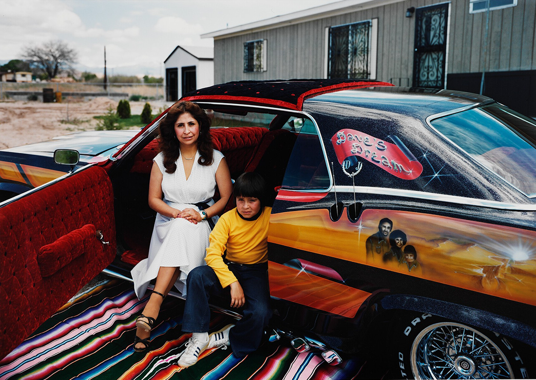 Person in a white dress and a young child sitting in a detailed car with a rug on the ground and a building in the background