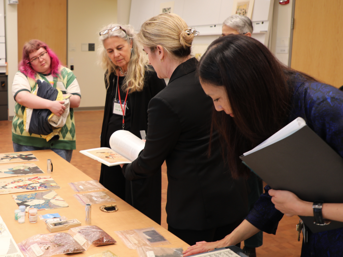 Five people standing over a table looking at colorful prints with one person showing images from a book
