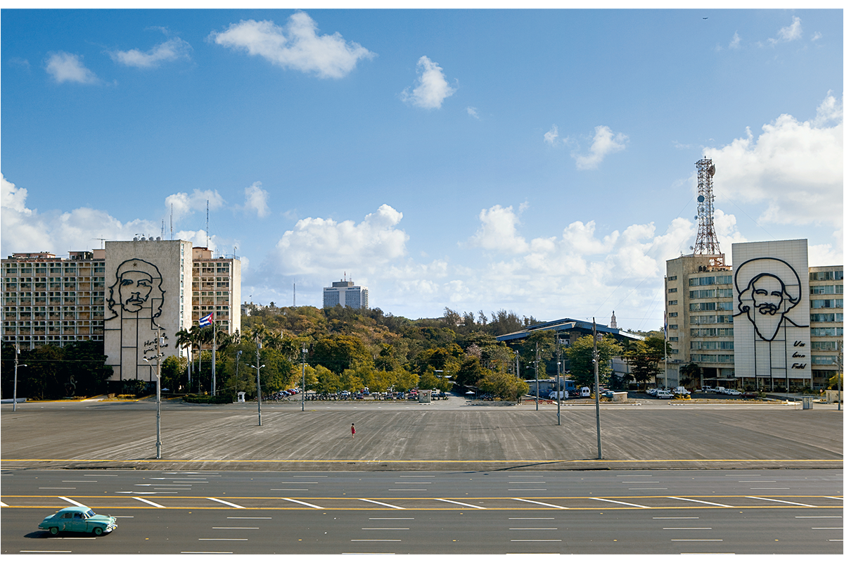 Streetscape with a baby blue old car in foreground with street in foreground, then an empty plaza and then buildings in background, two that have silhouettes of men on them