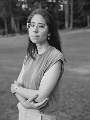 Black and white photo of a person with long hair, glasses, short sleeve shirt with arms crossed at waist