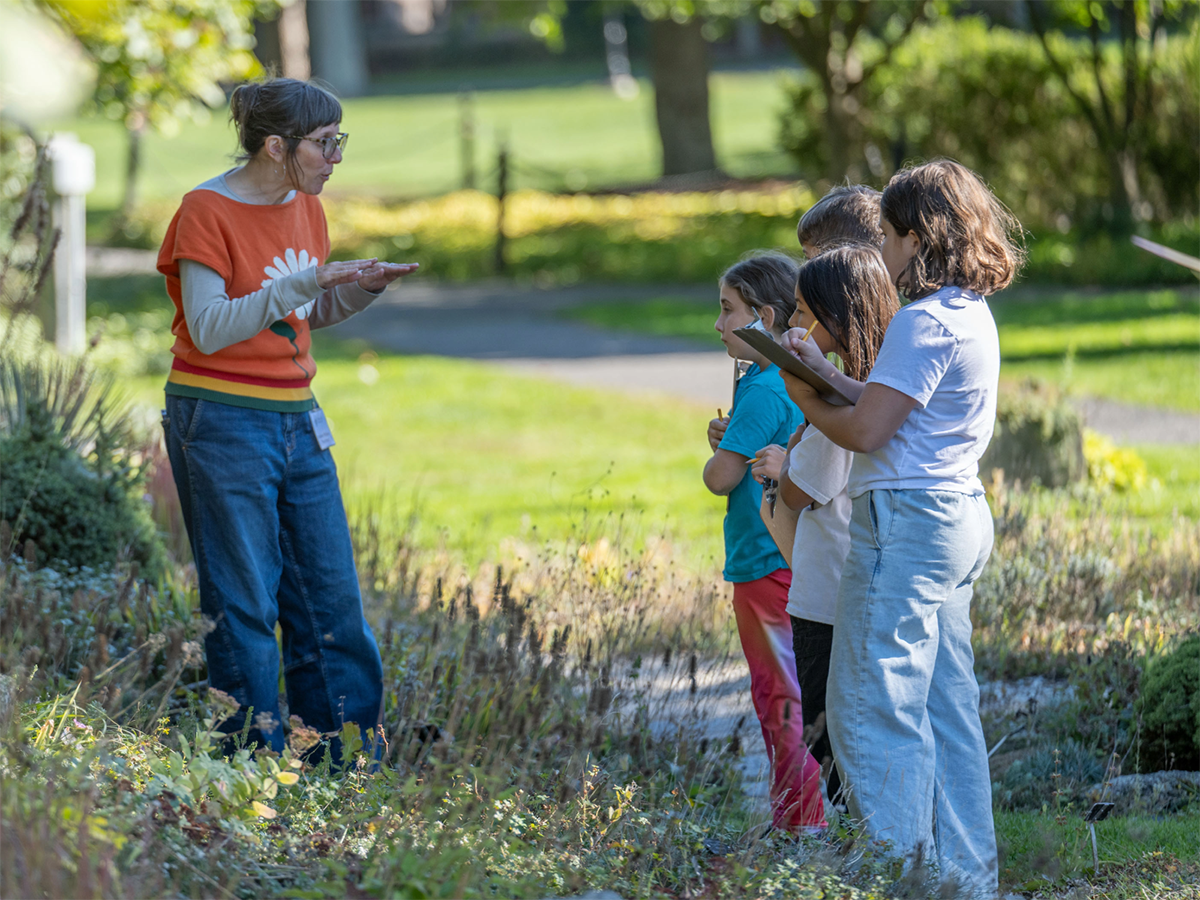 An adult outdoors explaining something to three kids with clip boards and one taking notes
