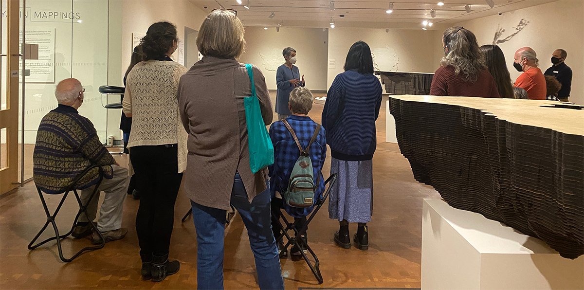 "Group of adults in Mappings exhibition facing woman presenting"