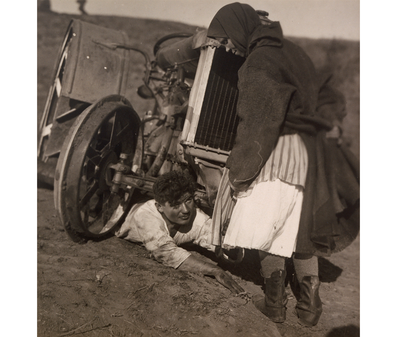 woman standing and man on the ground, under a wheel in a pit of dirt