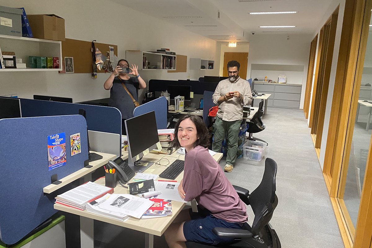 "Kamala GhaneaBassiri sitting at their desk at the MCA with their parents in the background"