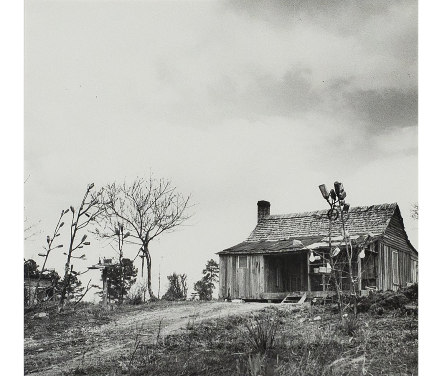 black and white photograph of a house with trees in the yard in front of it, some real trees and some trees constructed out of pieces of metal and branches