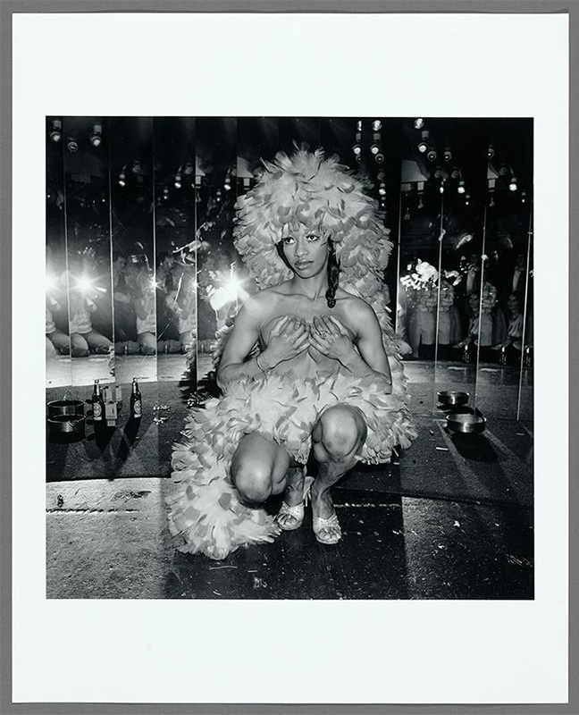 "black woman crouching in front of mirrors wearing a feathered headdress and high heels covering her breasts with her hands. People reflected in the mirrors"