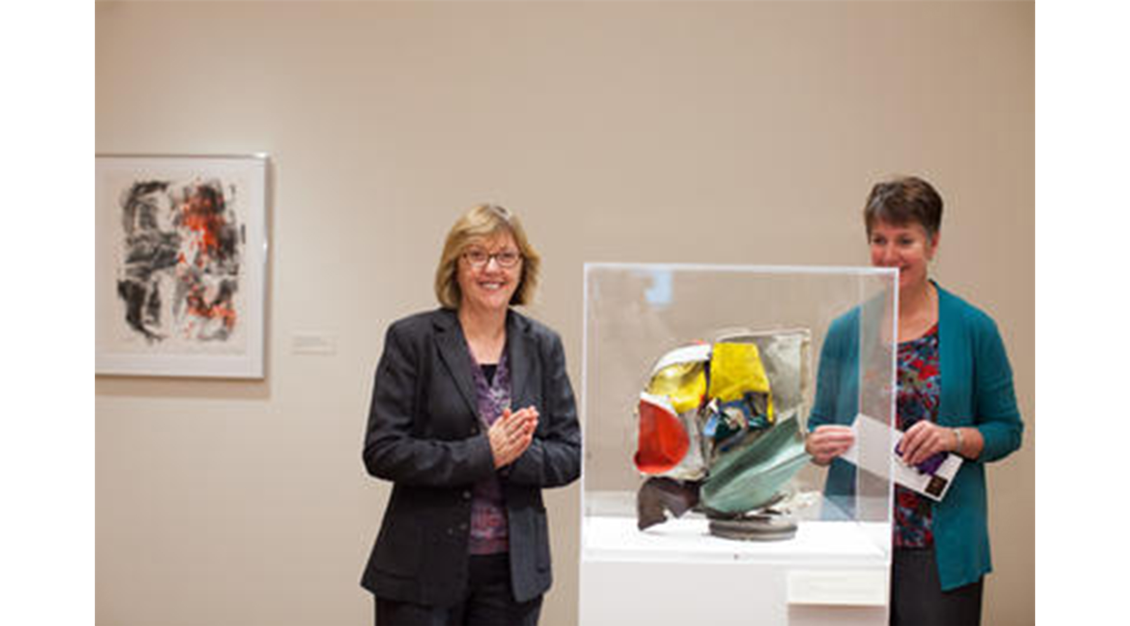 two women stand in an art gallery, looking at a glass case with a blue, yellow, and red sculpture inside of it. abstract painting on the wall behind them
