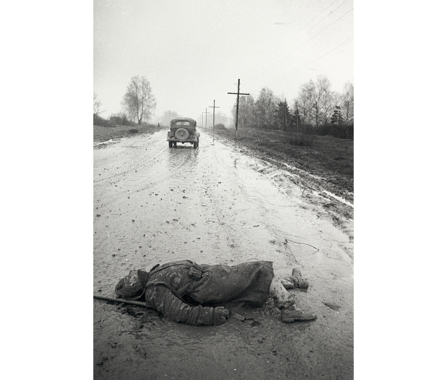soldier lying on the ground, covered in mud, in the middle of a road with one car driving on it