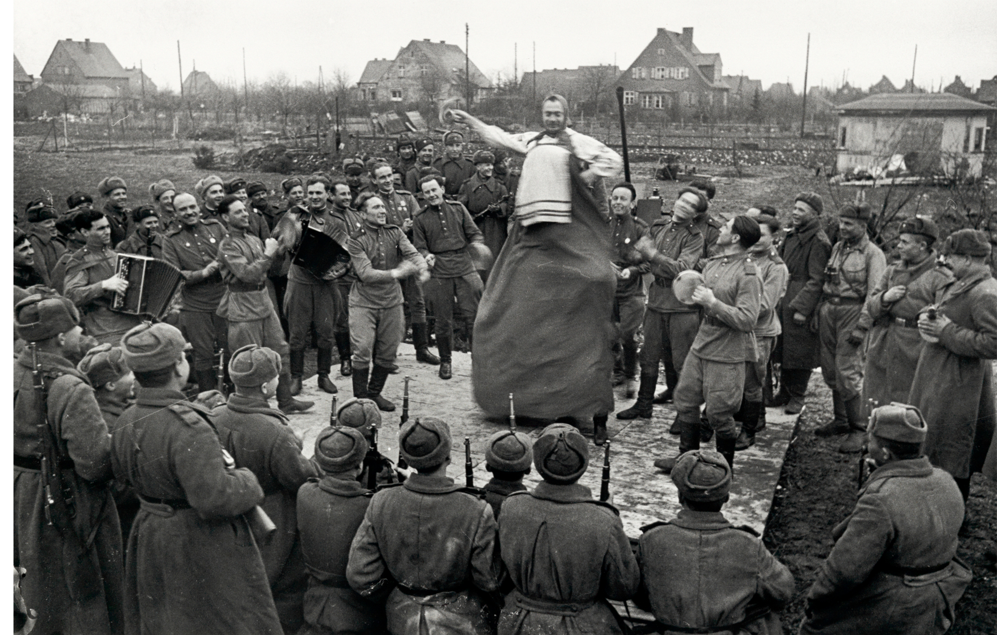 group of soldiers standing in a circle around a man walking on stilts; soldiers clap and cheer