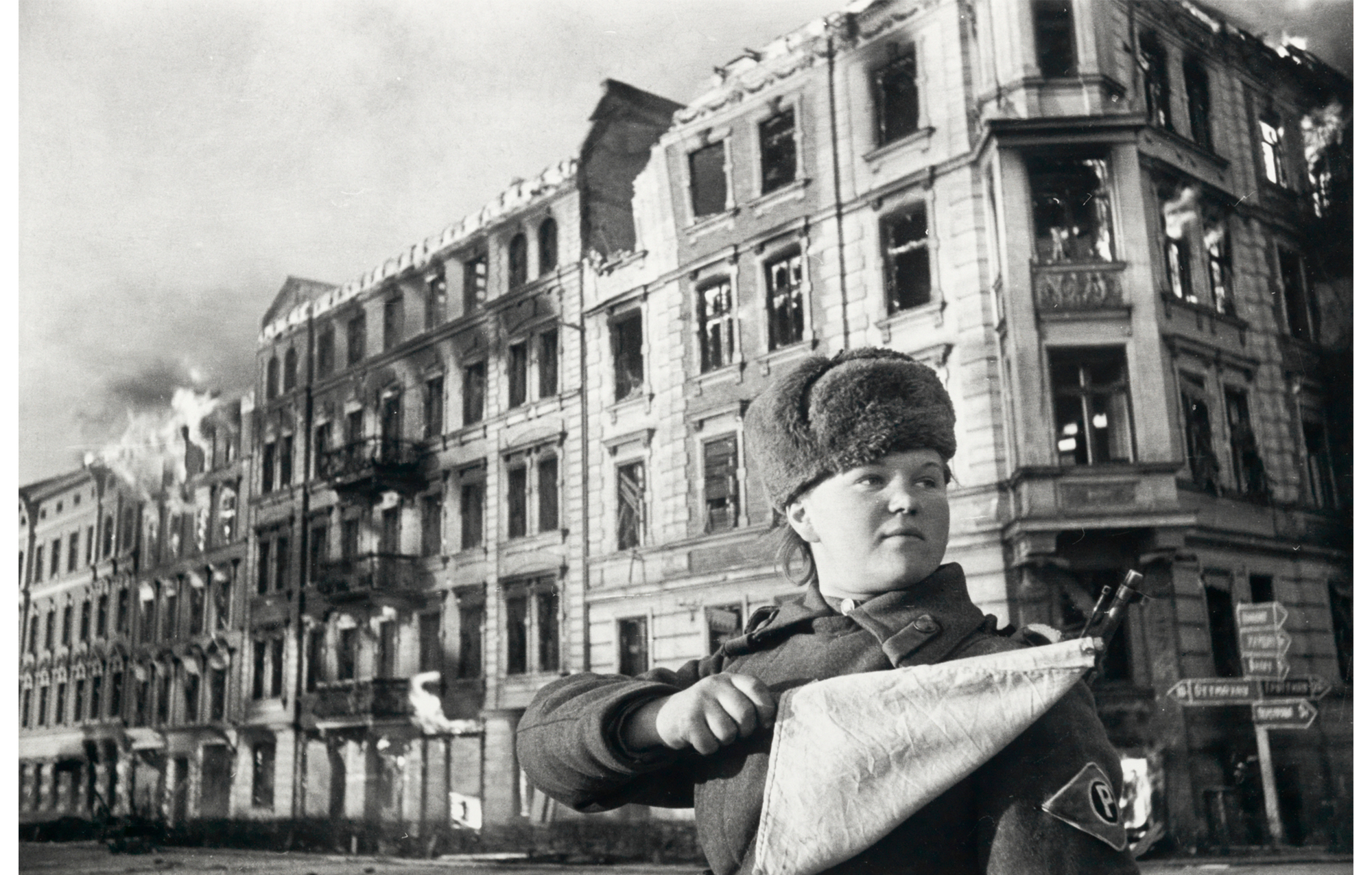female soldier wearing fur cap stands in front of a large building whose edge appears to have caught on fire