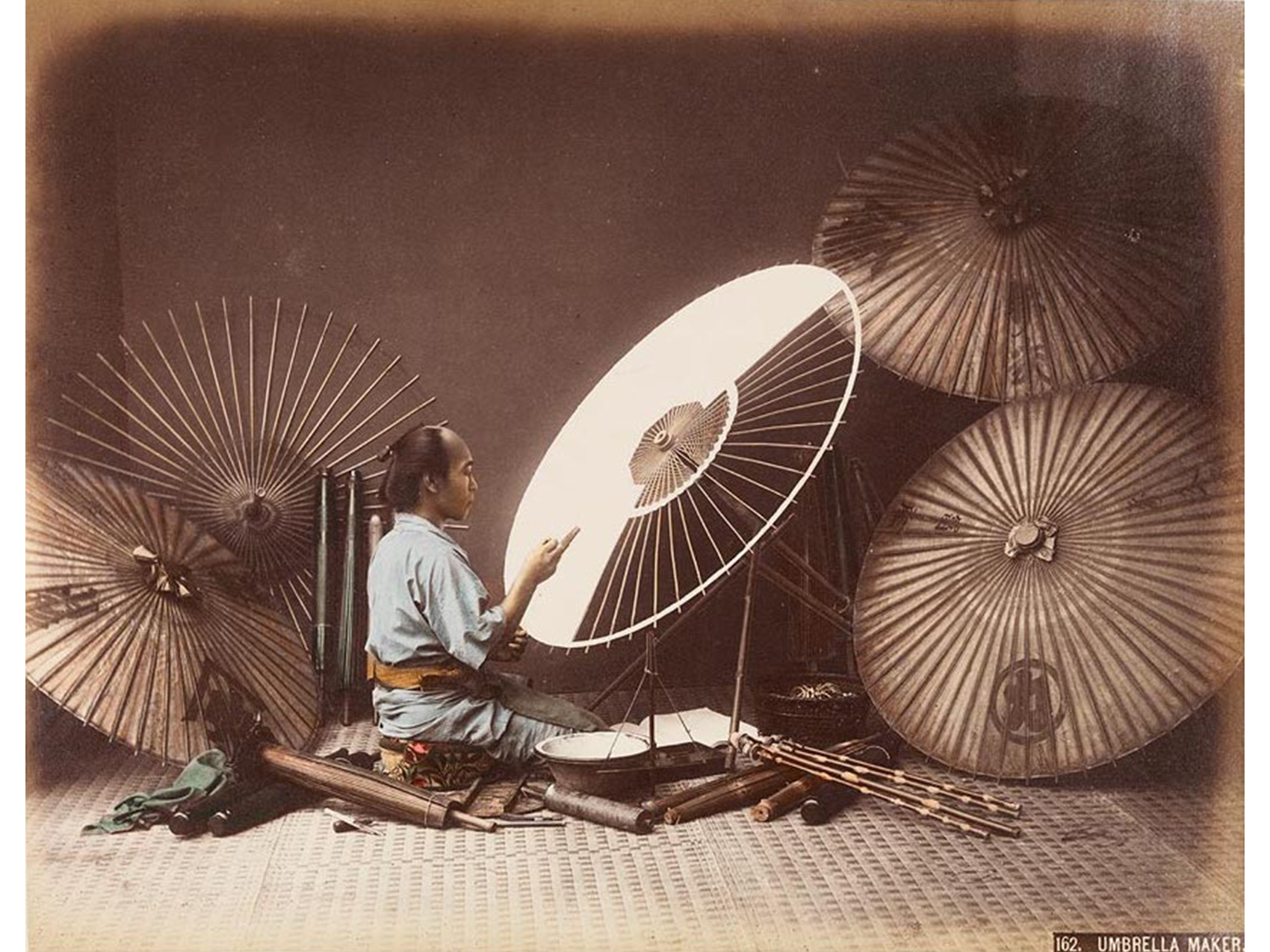 interior, man in green robe seated on woven mat working on a paper umbrella with three finished umbrellas and one unfinished open around him, several more scattered around him