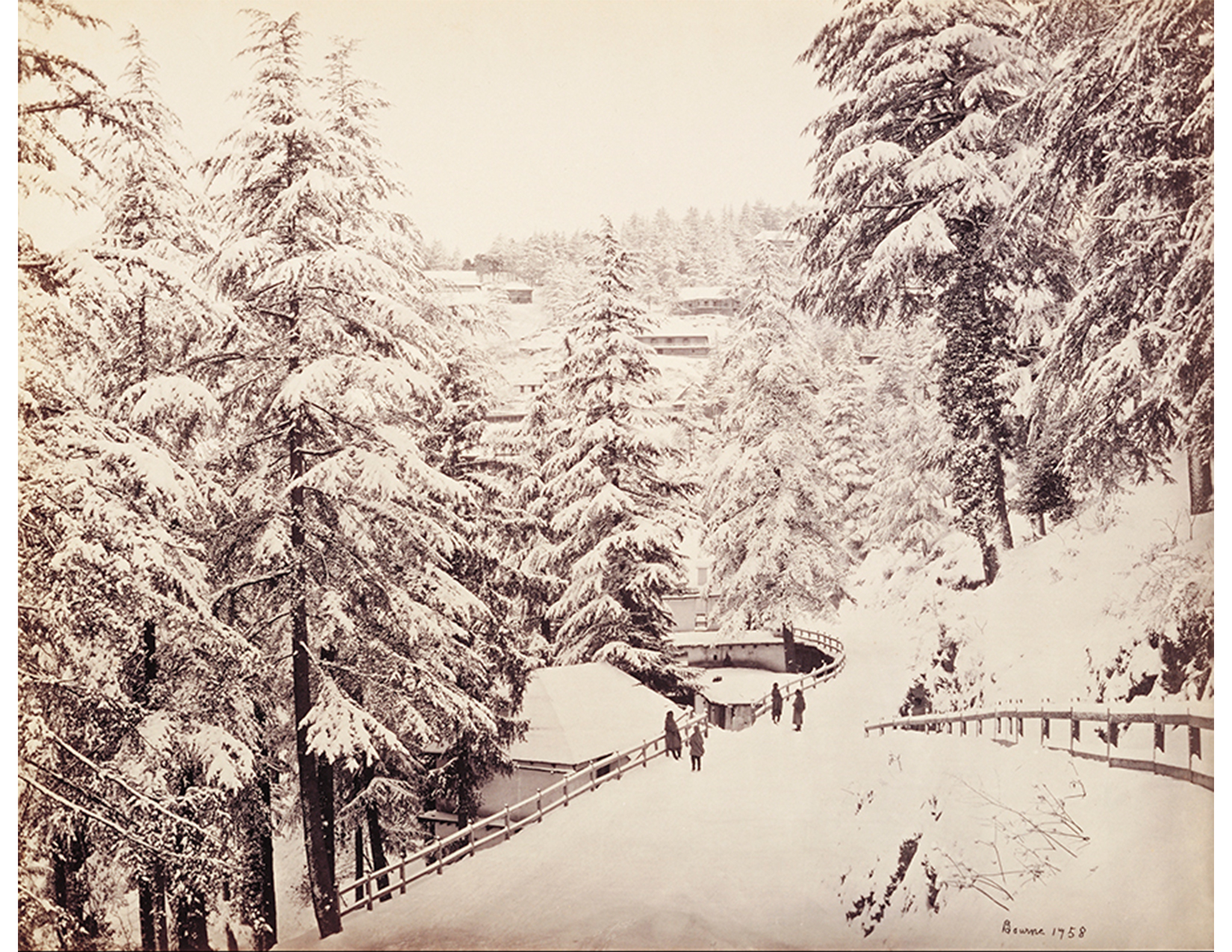 black and white image of four people walking on a snowy path lined with trees. in the background, a mountainside with buildings on it