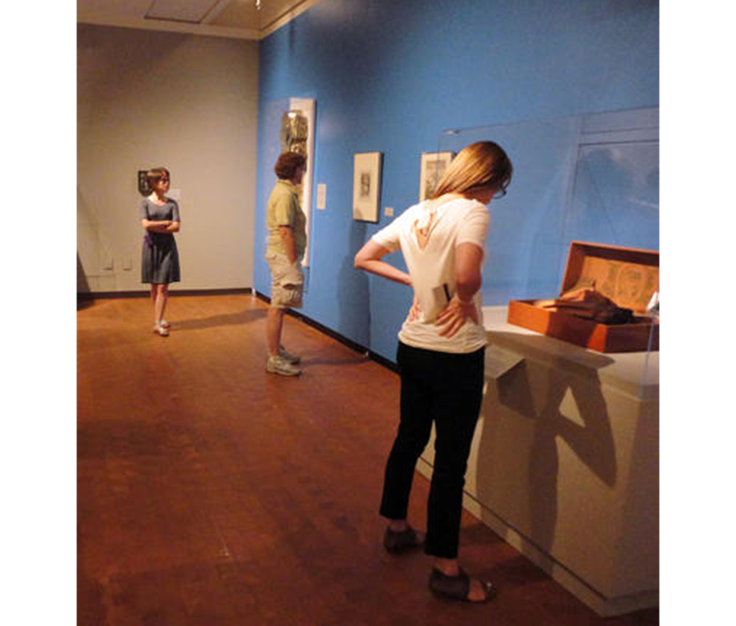 three people stand in an art gallery, looking at a wall painted blue with paintings and prints displayed and a display cabinet on the ground