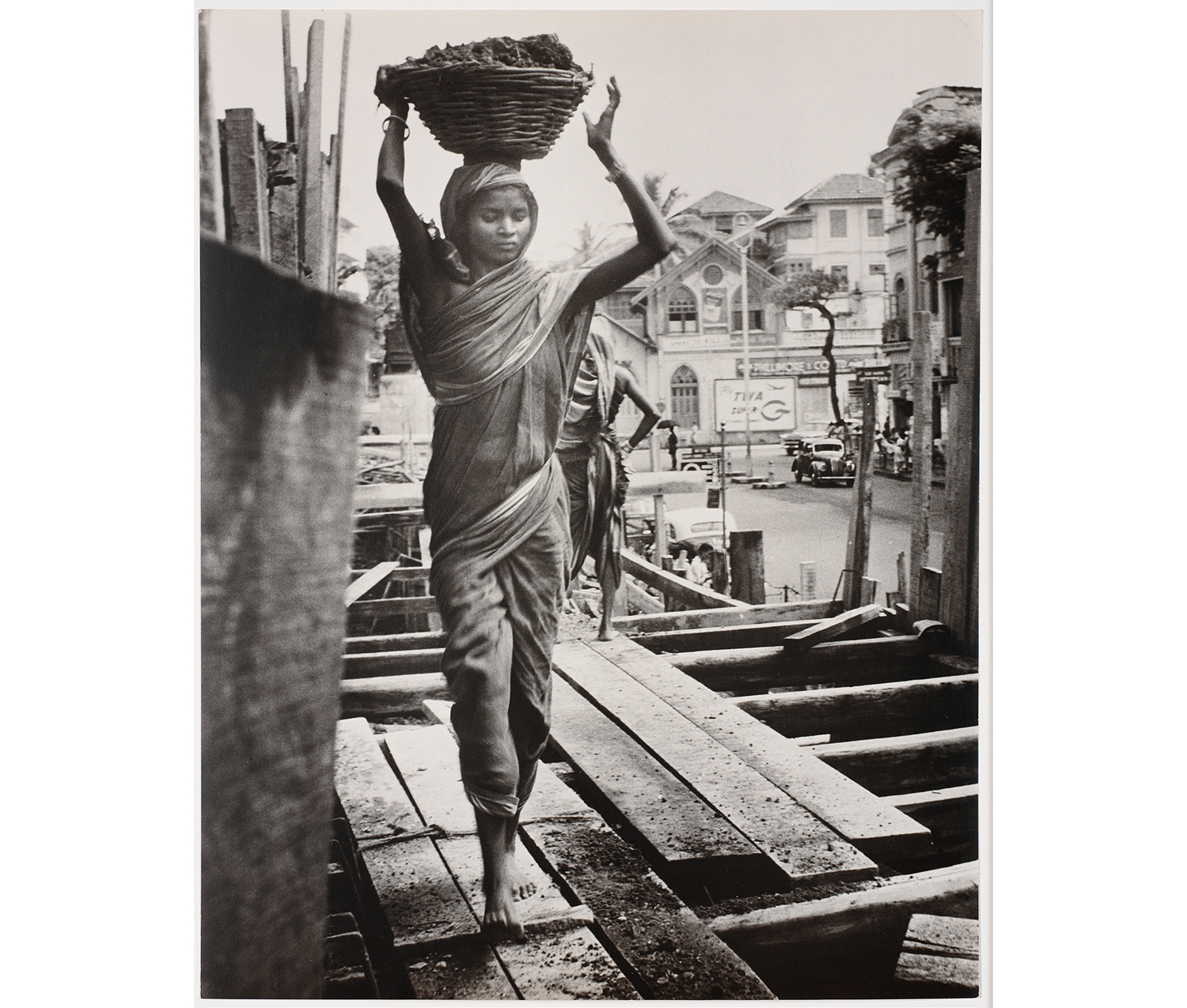 young person walking on a construction site, carrying a basket on their head