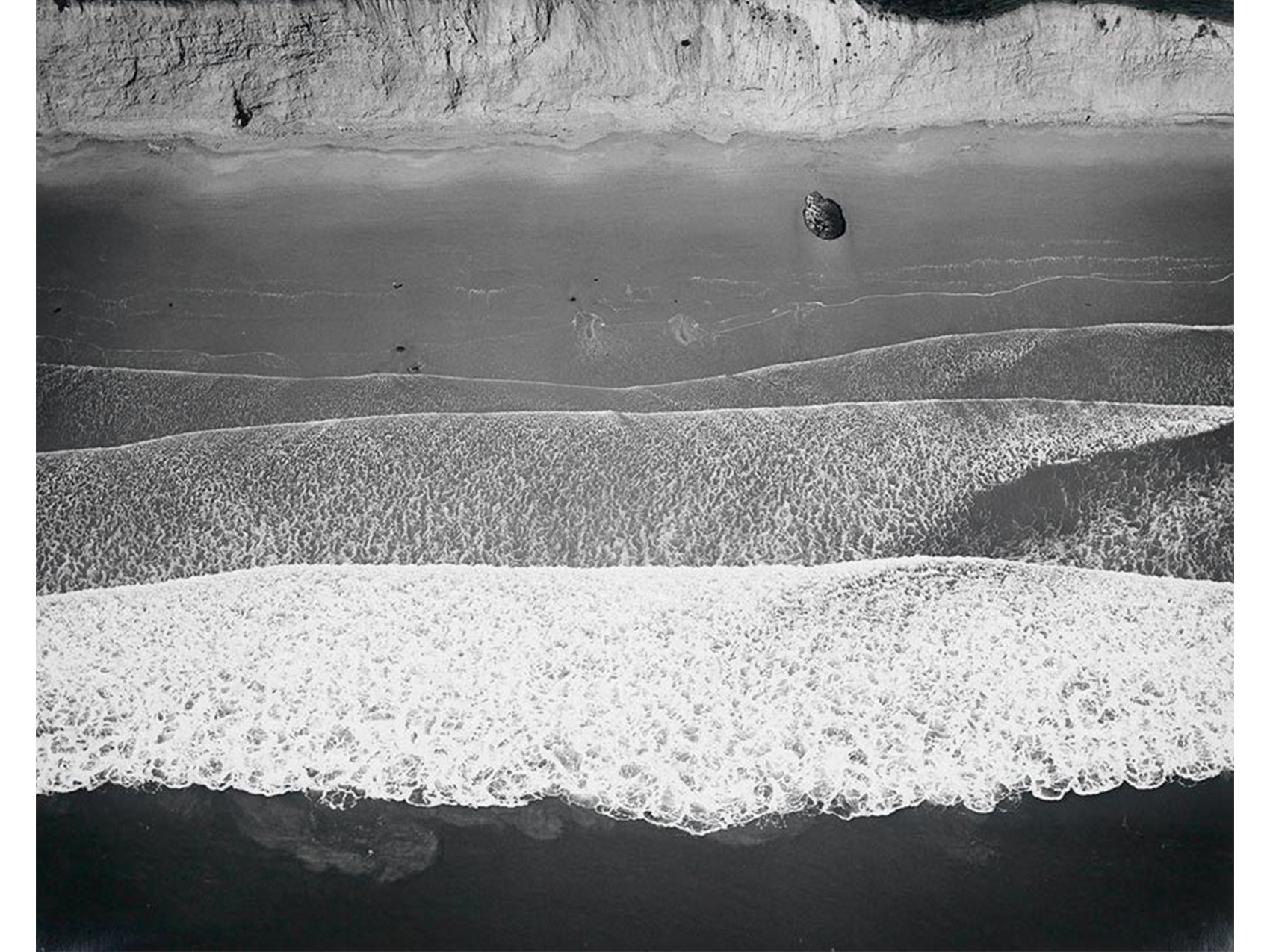 aerial view of shore with water and white foamy waves in lower image, beach and ridge of hill in upper half
