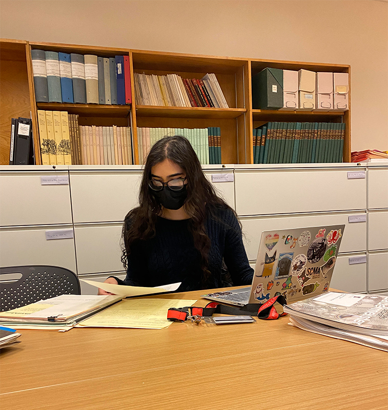 "Brooklyn sitting at her desk reading files with her open computer next to her"