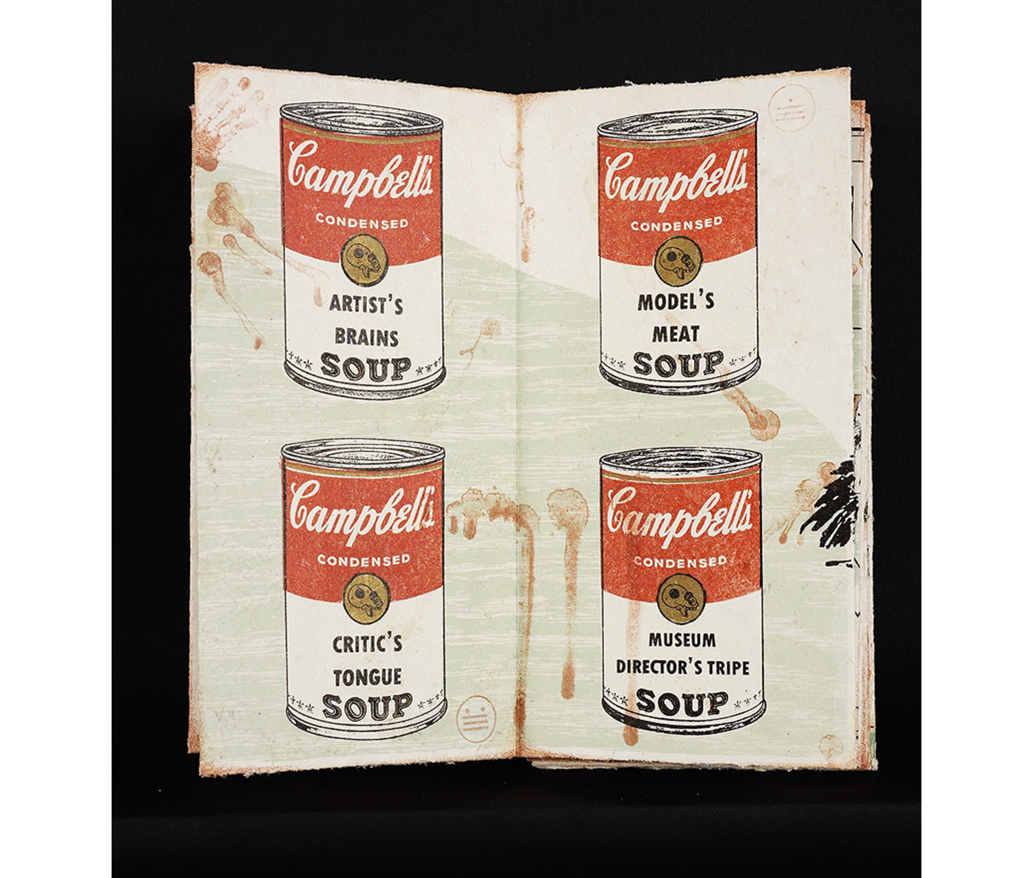 An open book displaying prints of four cans of Campbell’s Soup. The can on the top left says “Artist’s Brains Soup.” The can on the top right says “Model’s Meat Soup.” The can on the bottom left says “Critic’s Tongue Soup.” The can on the bottom right says “Museum Director’s Tripe Soup.”