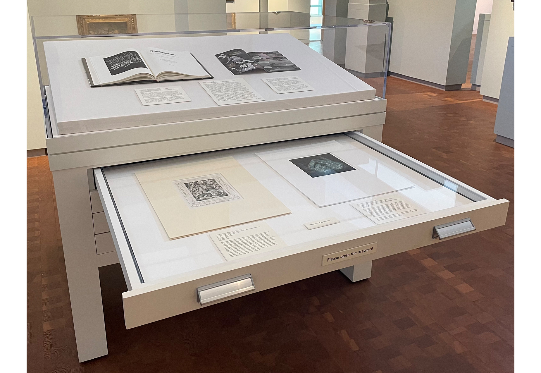 art gallery with display cabinet in the center, two open books on top of display cabinet and one drawer open to reveal two prints