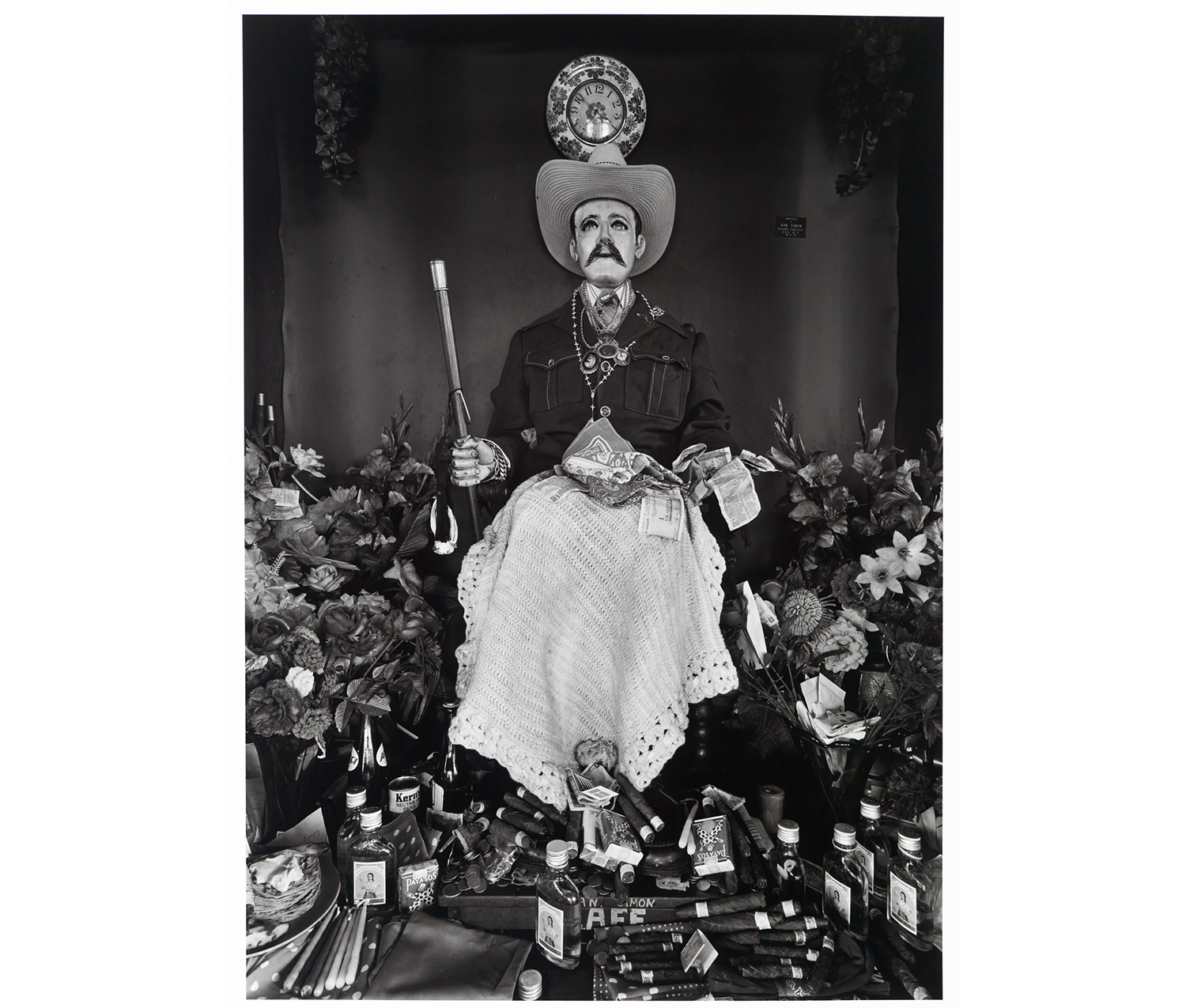 carved figure of a man with mustache, cowboy hat, coat, rosary around his neck, fake flowers around him, a floral clock over his head, cigars, alcohol, bandanas and money scattered around his feet and over his lap