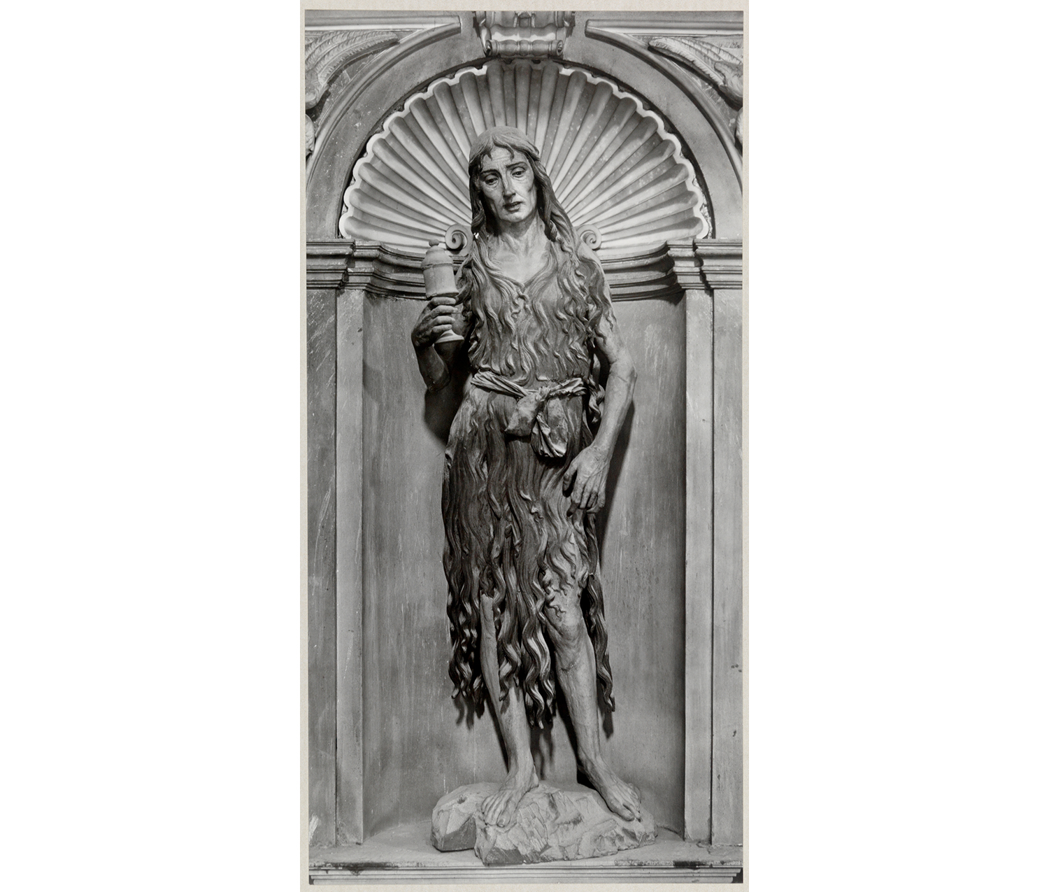 sculpture of a woman wearing a dress and holding a candle in her proper right hand, standing underneath an arch