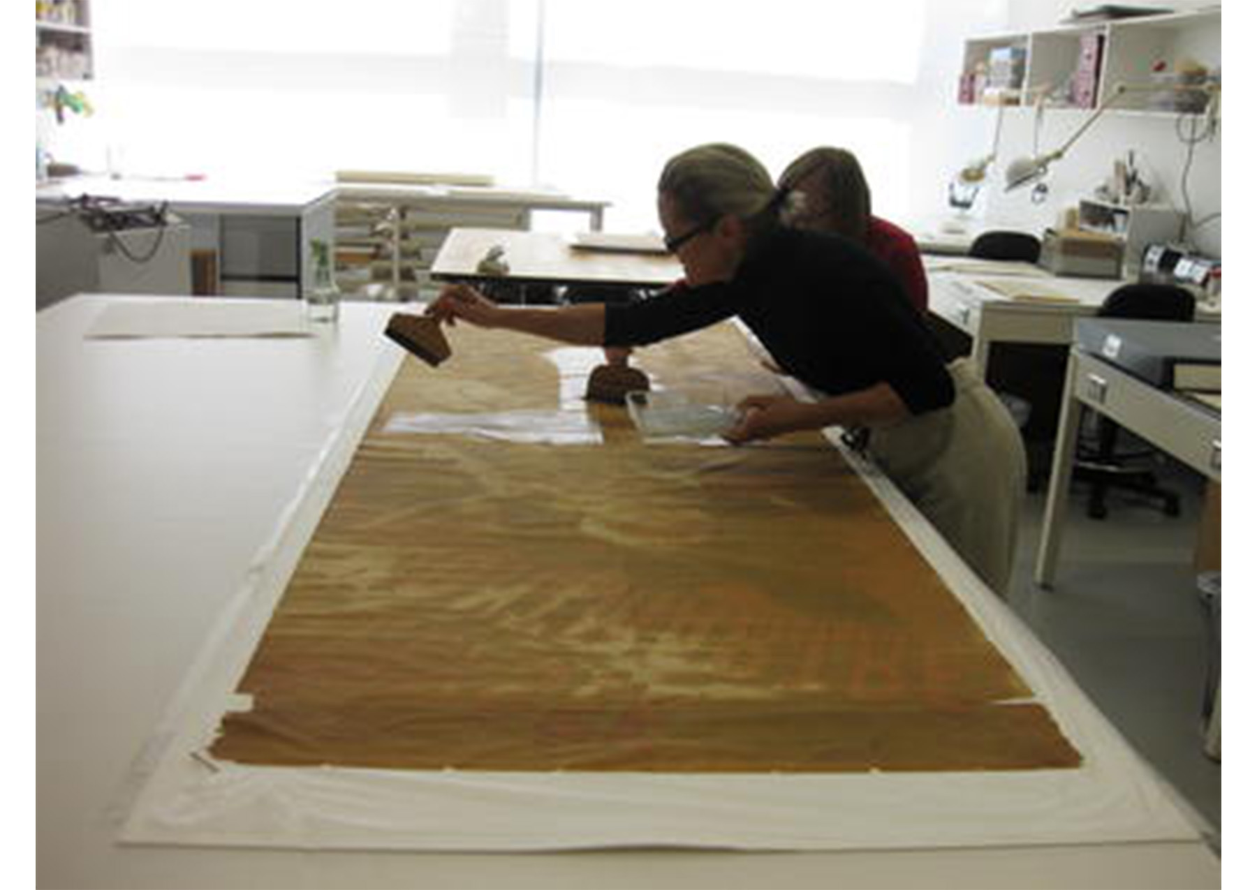 poster spread across a white table; two woman apply paste to the poster with brushes